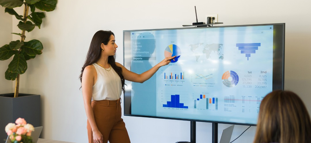 A person stands in front of a large display screen pointing at a pie chart among various other charts and graphs during a presentation.